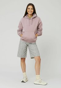 Hoodie violet clair avec poche kangourou, associé à un short imprimé léopard gris et des baskets beiges. Modèle se tenant avec les mains dans les poches.