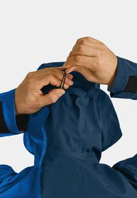 Blauer wasserdichter Jacke mit einer Kordelzugkapuze, sichtbaren Nähten und einem strukturierten Innenfutter. Hände ziehen den Kordelzug hinten an.