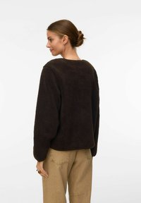 Sieviete ar matiem sasietiem kūkā, tērpusies melnā flīsa jaku un bēšās biksēs, nedaudz pagriezusies uz sāniem pret vienkāršu baltu fonu.