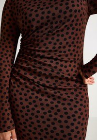 Brun kjole med sorte prikker, lange ærmer og indsamlet tekstur på den ene side. Stoffet ser glat og tætsiddende ud.