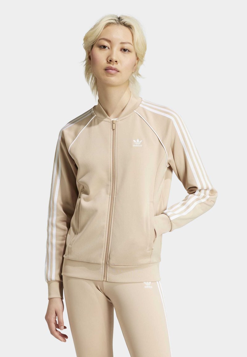 Chaqueta beige con cremallera y acentos de tres rayas blancas en las mangas. Confeccionada con un tejido suave, tiene un diseño ajustado y cuello alto.