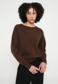 Καφέ, υπερμεγέθη πλεκτό πουλόβερ με ριμπ λαιμόκοψη και μανσέτες, με υφή υφάσματος. Συνδυάζεται με σκούρο παντελόνι.