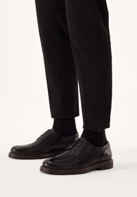 Pantofi negri din piele, cu un finisaj neted, vârful rotund și design cu șireturi, asortați cu pantaloni și șosete negre. Accente de brad pe talpa exterioară.