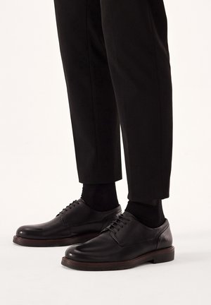 Scarpe nere in pelle con una finitura liscia, punta arrotondata e design con lacci, abbinate a pantaloni neri e calze. Accentazione con suola marrone.