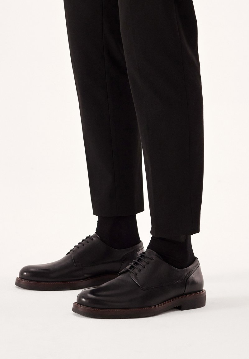 Pantofi negri din piele, cu un finisaj neted, vârful rotund și design cu șireturi, asortați cu pantaloni și șosete negre. Accente de brad pe talpa exterioară.