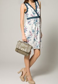 Robe florale avec une base blanche, des motifs bleus et roses, un col à volants, assortie à un sac à main kaki et des escarpins beiges.