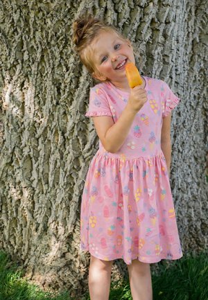 Junges Mädchen in einem rosa Kleid mit Eis und bunten Mustern steht lächelnd an einem großen Baumstamm im Freien.