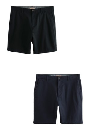 Junger Mann steht, trägt ein weißes oversized T-Shirt, schwarze Shorts und weiße Sneaker mit schwarzen Streifen und Gummisohlen.