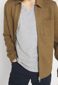 Mann in einer offenen braunen Zip-Jacke über einem hellgrauen V-Neck-Shirt und dunklen Navy-Hosen, mit einer Hand in der Tasche.