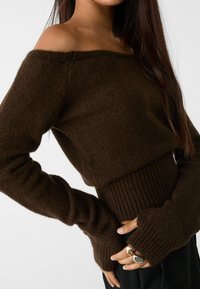 Femme portant un pull en tricot brun foncé à épaules dénudées avec des manches longues et des ouvertures pour les pouces, associé à un pantalon noir et des bagues aux doigts.