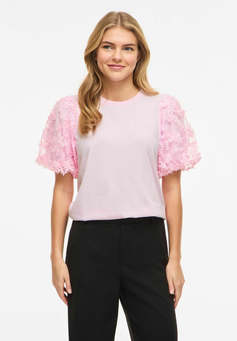 Młoda kobieta o falowanych blond włosach, ubrana w jasnoróżowy top z teksturowanymi bufkami na rękawach oraz czarne spodnie, z delikatnym uśmiechem, na białym tle.