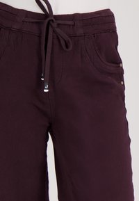 Donkerpaarse broek met zijzakken en een strikceintuur, waarin gedetailleerd stikwerk en de textuur van de stof zichtbaar zijn.