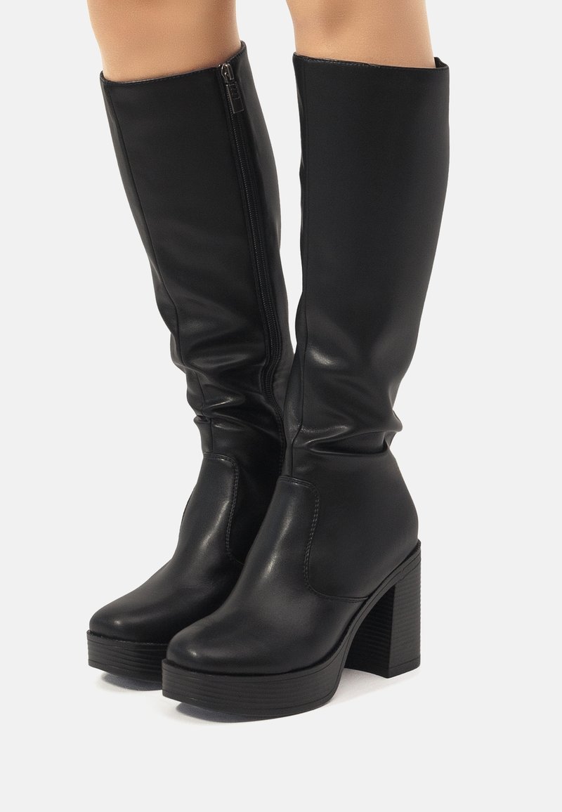 Bottes en cuir noires montant jusqu'aux genoux avec talons blocs et fermetures éclair latérales, portées sur des jambes nues, sur un fond uni.