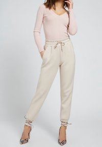 Haut à manches longues côtelé rose clair avec col en V ; pantalon de survêtement beige effilé avec taille à cordon et bas élastiqués ; chaussures à talons hauts à motif.