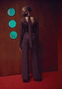 Mono de encaje en un profundo morado con mangas largas y espalda descubierta, que presenta un detalle atado en el cuello. Ajustado en la parte superior, con pantalones acampanados en la parte inferior.