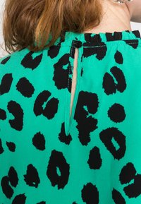 Top de tecido verde com padrão de leopardo preto, com um fecho em forma de olho na parte de trás preso por um botão. Textura suave, design casual.