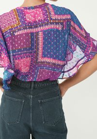 Blusa colorata e leggera con un design patchwork in viola, rosa e blu; tessuto trasparente con vari motivi geometrici. Si abbina a jeans scuri a vita alta.