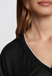 Zwarte top met een glanzende textuur, met een V-halslijn omzoomd met zilveren rand. Glad materiaal met een comfortabele pasvorm rond de hals.