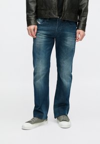 Jeans di denim blu con lavaggio sbiadito e design a gamba dritta, abbinati a scarpe in tela grigie e giacca di pelle nera.