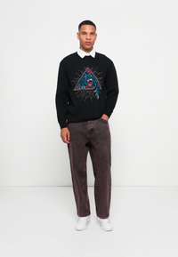 Man wearing black sweater with blue roaring panther graphic over white collared shirt, purple washed jeans, and white sneakers, standing against white wall.