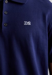 Polo de color azul marino hecho de tejido texturizado, con una tapeta de dos botones y un logo bordado "D2" en blanco en el pecho.
