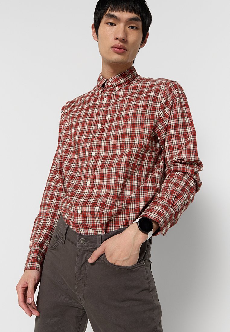 Camisa de cuadros roja y crema con cuello. Mangas largas con puños abotonados, llevada con pantalones grises. Reloj en la muñeca izquierda.