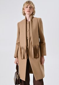 Cappotto color cammello con una silhouette strutturata, dotato di un colletto alto, chiusure a bottone e dettagli frangiati all'orlo.