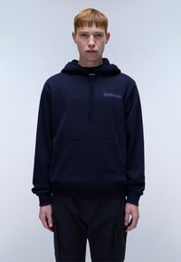 Marineblauwe hoodie met een zak aan de voorkant, een trekkoord kap, zachte stof en geborduurd logo op de borst. Model staat tegen een lichte achtergrond.
