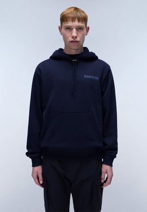 Marineblauwe hoodie met een zak aan de voorkant, een trekkoord kap, zachte stof en geborduurd logo op de borst. Model staat tegen een lichte achtergrond.
