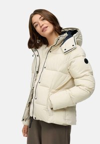Femme portant une doudoune beige clair avec capuche et zipper, se tenant avec une main dans la poche devant un fond uni.