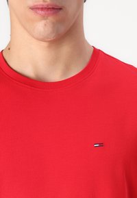 Röd bomullströja med rund halsringning och korta ärmar. Har en liten broderad logotyp på vänster bröstområde.