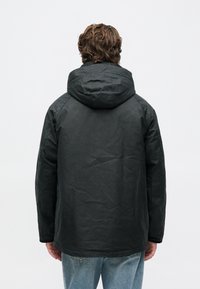 Bărbat cu păr ondulat maroniu care poartă o jachetă cu glugă neagră și blugi ușor albaștri, întors cu spatele pe un fundal alb simplu.