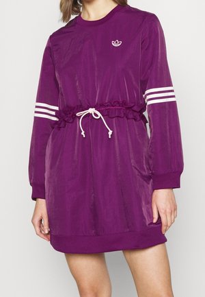 Kobieta w fioletowej sukience z długim rękawem, z białymi paskami na rękawach, białym ściągaczem w talii i małym białym logo Adidas na piersi.