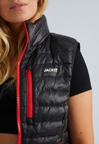 Schwarze gesteppte Weste mit roten Reißverschlüssen, ausgestattet mit einem Stehkragen und dem Logo "JACKIT" in Weiß auf der Vorderseite. Leichter Stoff.