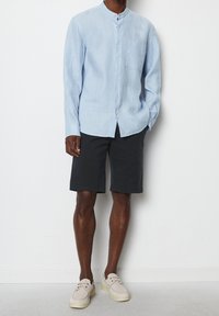 Vaaleansininen pellavapaita pitkällä hihalla, jossa on mandariinikaulus ja etutasku. Yhdistetty mustiin shortseihin ja beigenvärisiin nauhakenkiin.