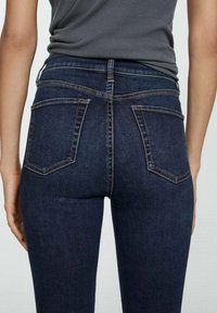 Jean en denim bleu foncé avec une coupe ajustée, taille haute et deux poches arrière avec couture contrastée. Texture lisse et style classique.
