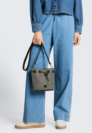 Persoon draagt lichtblauwe jeans met wijde pijpen, spijkerjack, witte sneakers, en houdt een kleine zwart-wit geweven emmertas met zwarte bandjes vast.