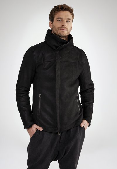 Schwarze Wildlederjacke mit hohem Kragen, asymmetrischer Vorderseite und zwei seitlichen Reißverschlusstaschen; glatte Textur, taillierte Passform und modernes Design.