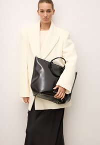 Čierna kožená taška s lesklým povrchom, dvojitými držadlami a kovovými akcentmi, ktorá je držaná vedľa modelky oblečenej v bielom kabáte.