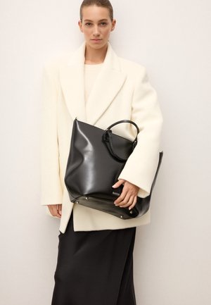 Črna usnjena torba z visokim sijajem, dvojni ročaji in kovinski detajli, ki jo drži model v belem plašču.