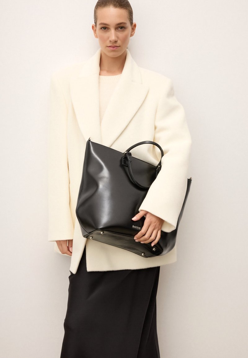 Čierna kožená taška s lesklým povrchom, dvojitými držadlami a kovovými akcentmi, ktorá je držaná vedľa modelky oblečenej v bielom kabáte.