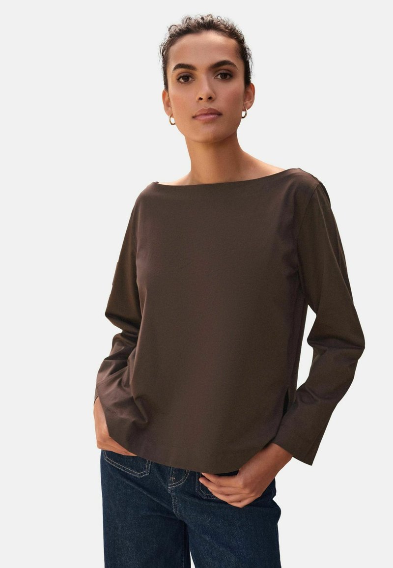 Brown long-sleeve top with a boat neckline, made of smooth fabric, featuring side slits. Paired with blue jeans.