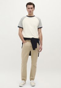 T-shirt à manches courtes beige et gris à manches raglan, pantalon beige, pull marine noué autour de la taille et baskets crème. Tenue simple et décontractée avec des lignes épurées.