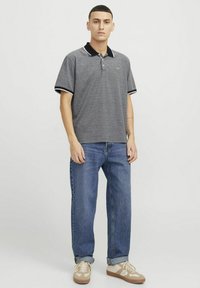 Pilkas polo marškinėliai su juodu apykakle ir rankogaliais, trumpomis rankovėmis; dėvimi su mėlynais džinsais ir šviesiai rudos spalvos sportbačiais. Audinys atrodo lygus ir kasdieniškas.