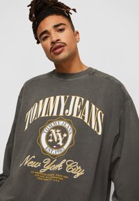 Felpa oversize grigio scuro con grafica "TOMMY JEANS", caratterizzata da accenti gialli e un logo circolare che recita "New York City" sotto.