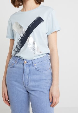 Henkilö, jolla on yllään vaaleansininen t-paita, jossa on musta ja hopeinen siveltimenveto X-kuvio, ja joka on työnnetty korkeavyötäröisiin vaaleisiin farkkuihin.