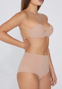 Nudová podprsenka bez ramínek s hladkým konturem, tvarovanými košíčky a průhledným pásem, v kombinaci s vysokými nudovými kalhotkami. Měkká, bezešvá tkanina.
