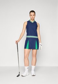 Vestido de golfe em azul-marinho com detalhes em verde, apresentando um decote em V e design sem mangas. Inclui uma saia plissada e uma faixa listrada na cintura. Luvas e sapatos de golfe brancos.