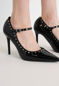 Zwarte gepatineerde leren hoge hakken met een puntige neus, voorzien van een bandje met een zilveren gesp en decoratieve zilveren studs langs de rand.