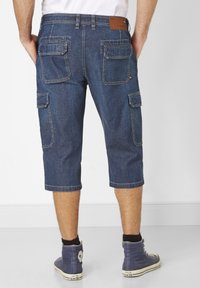 Denim cargo shorts in dark blue with a relaxed fit, knee-length, featuring multiple pockets and a stitched hem. Worn with blue high-top sneakers.